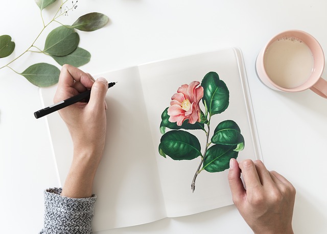 a hand holding a pen, poised over a journal with a blank page on one side and a drawing of a pink flower on the other